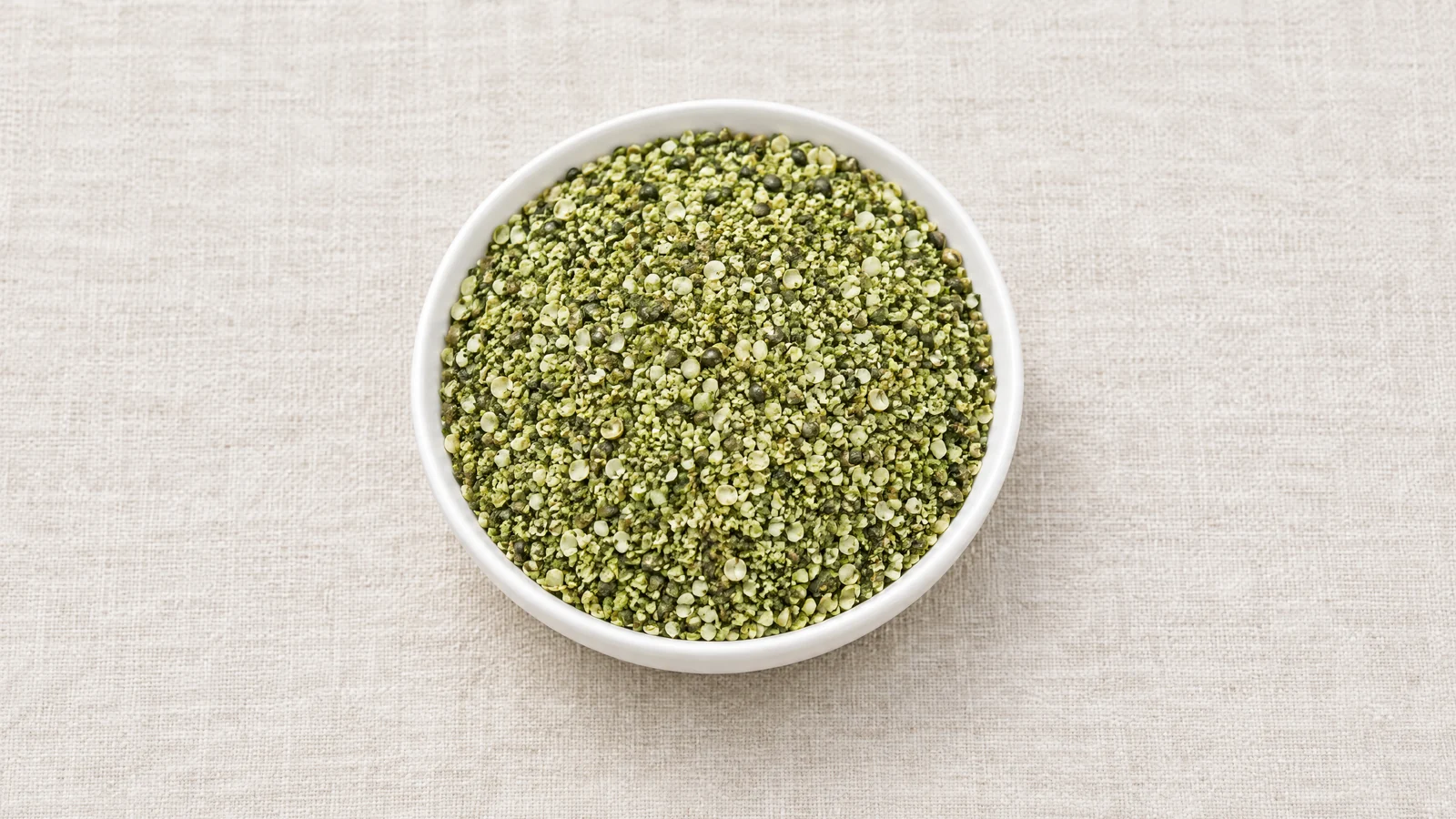 Granular smoothie hemp, small green kernel and hull fragments, in a white ceramic dish on linen